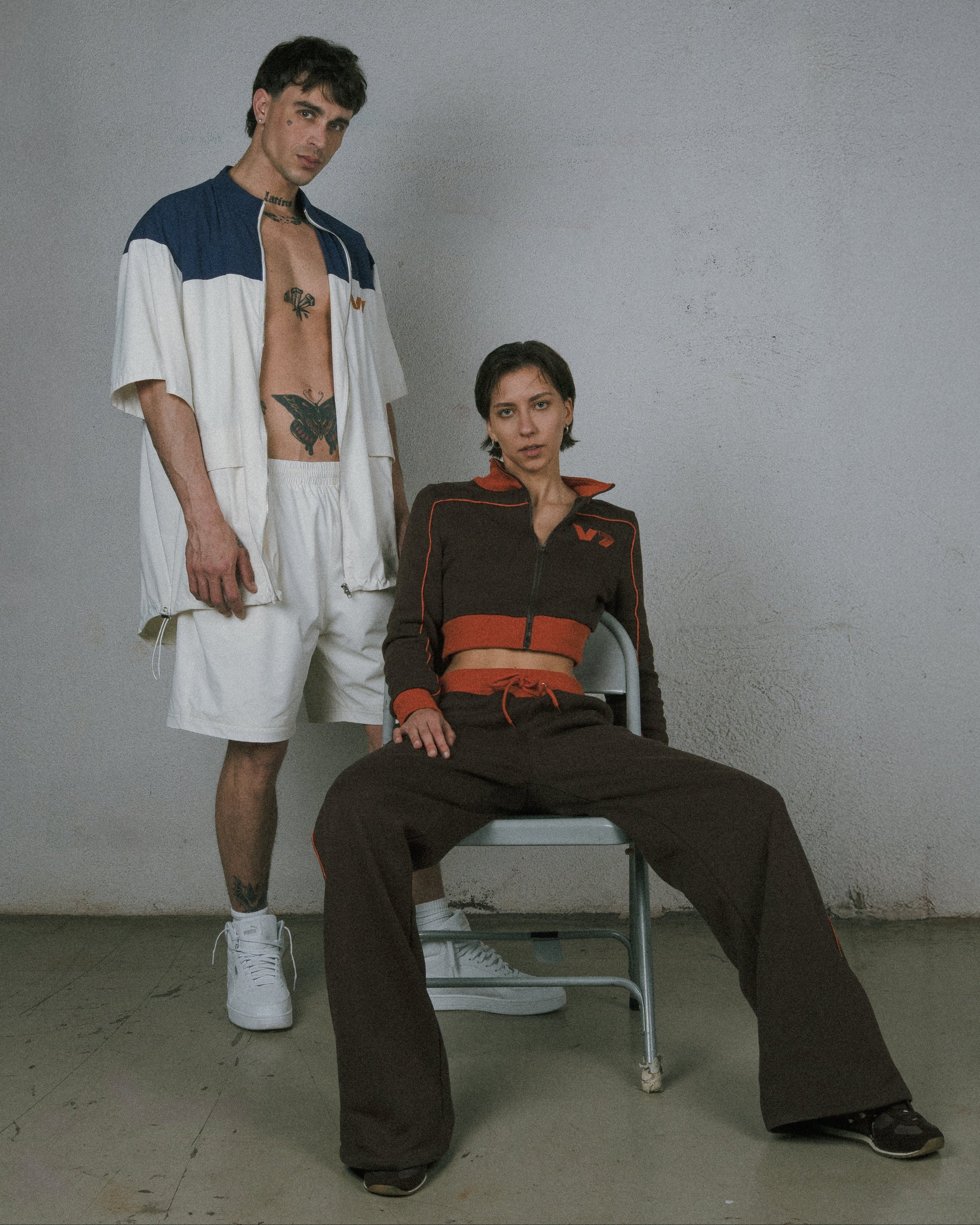 Two people posing against a plain wall, one standing and one sitting on a chair.
